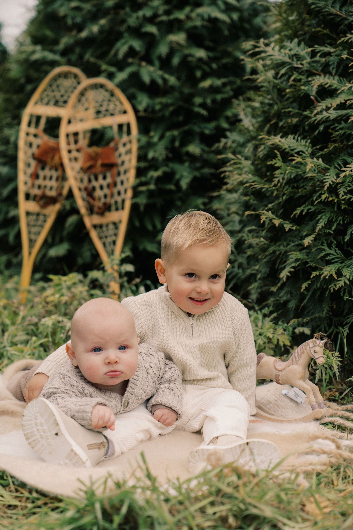 Two children in cozy cream sweaters lay on a blanket outdoors with vintage snowshoes visible in the background.