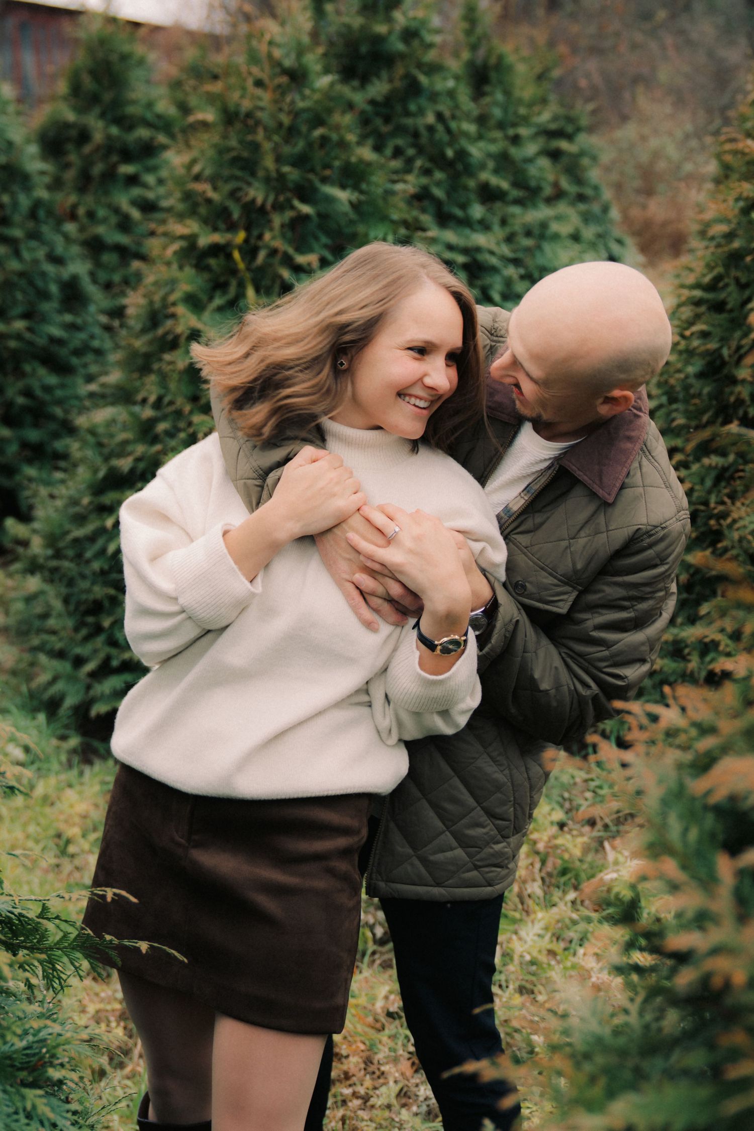 Couple sharing playful moments between evergreen trees at a Christmas tree farm.