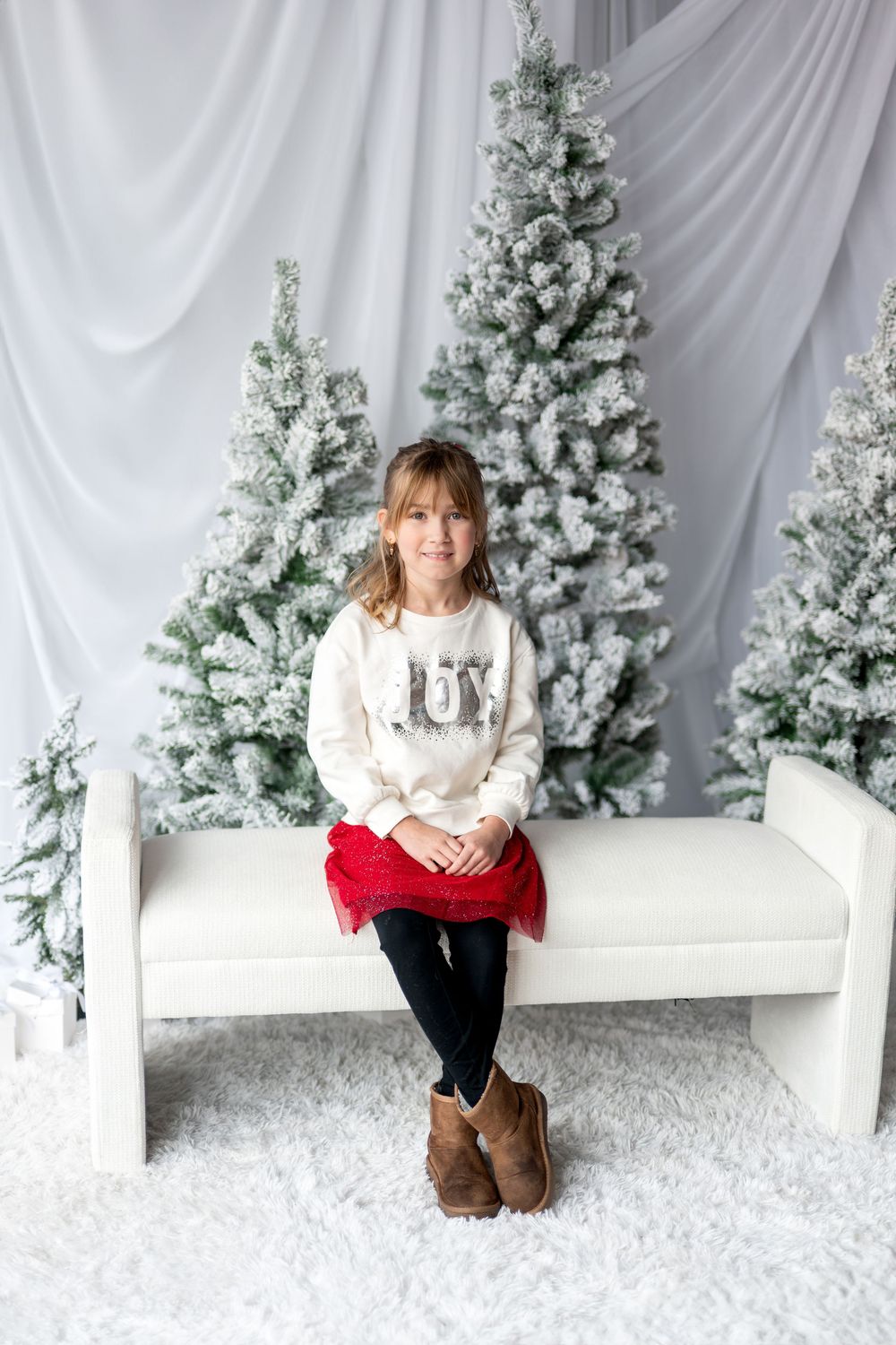 Young girl in JOY sweater and red skirt sits on white bench before snow-flocked Christmas trees.