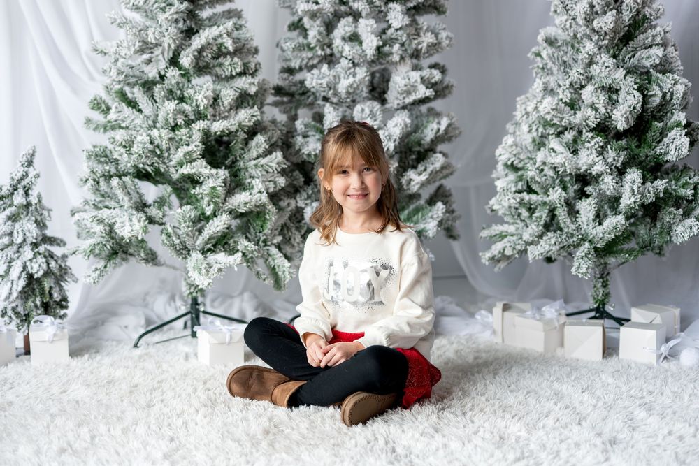 Young girl in white reindeer sweater sits cross-legged on fluffy rug before flocked Christmas trees.