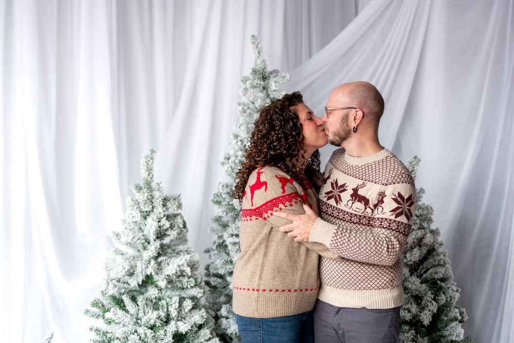 Couple kissing in matching Christmas sweaters with reindeer patterns by snow-flocked holiday trees.