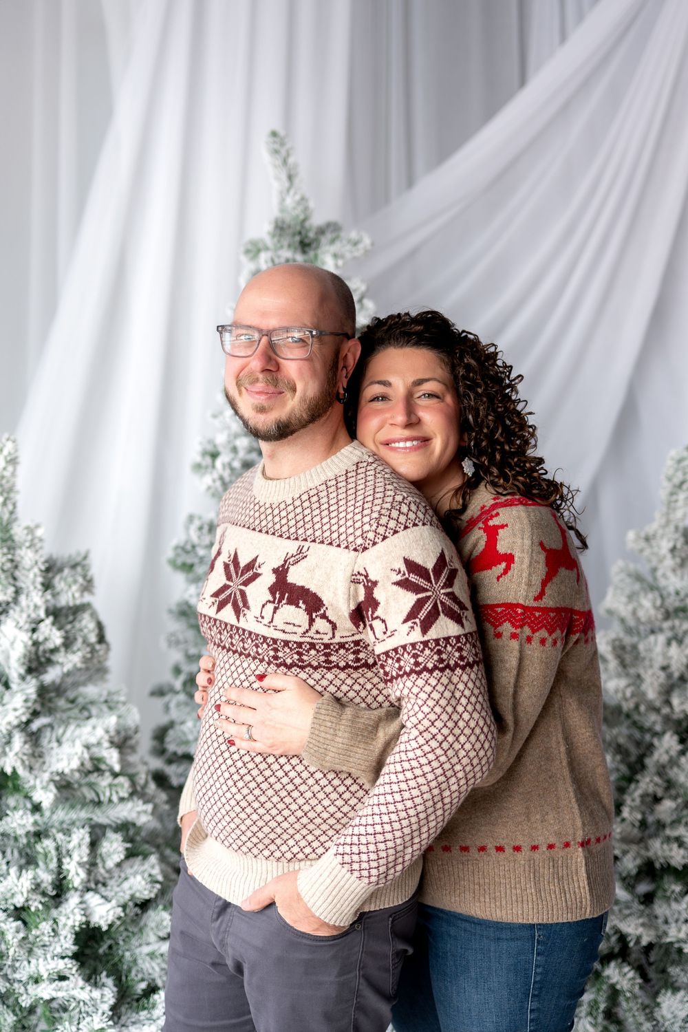 Happy couple embracing in coordinating Nordic reindeer sweaters in front of snowy Christmas trees.