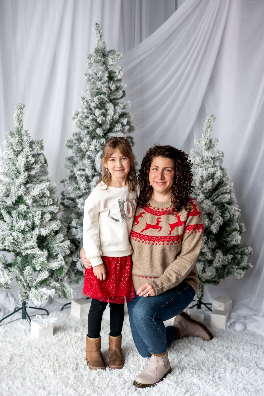 Mother and daughter in reindeer sweaters posing together beside snow-flocked Christmas trees.
