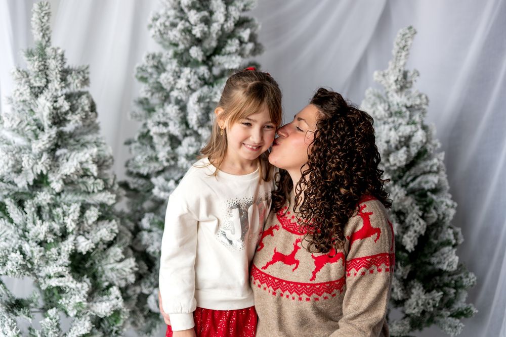 Mother kissing young daughter on cheek in front of snow-flocked Christmas trees in holiday sweaters.