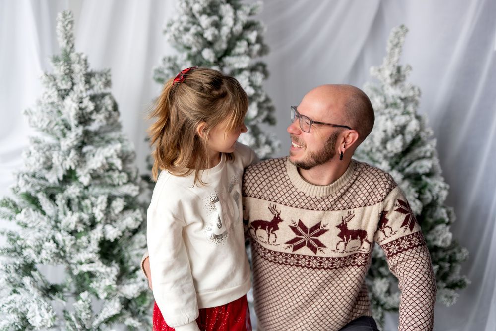 Father and daughter in matching reindeer sweaters smiling at each other by snowy Christmas trees.