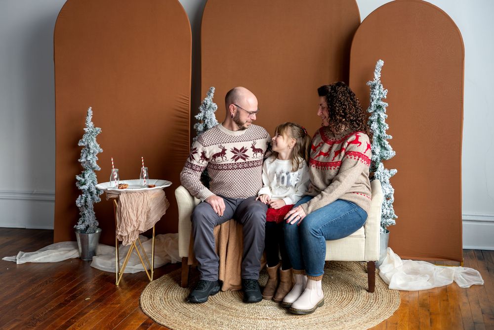 Family in matching reindeer sweaters sitting and chatting on a bench with Christmas trees behind.