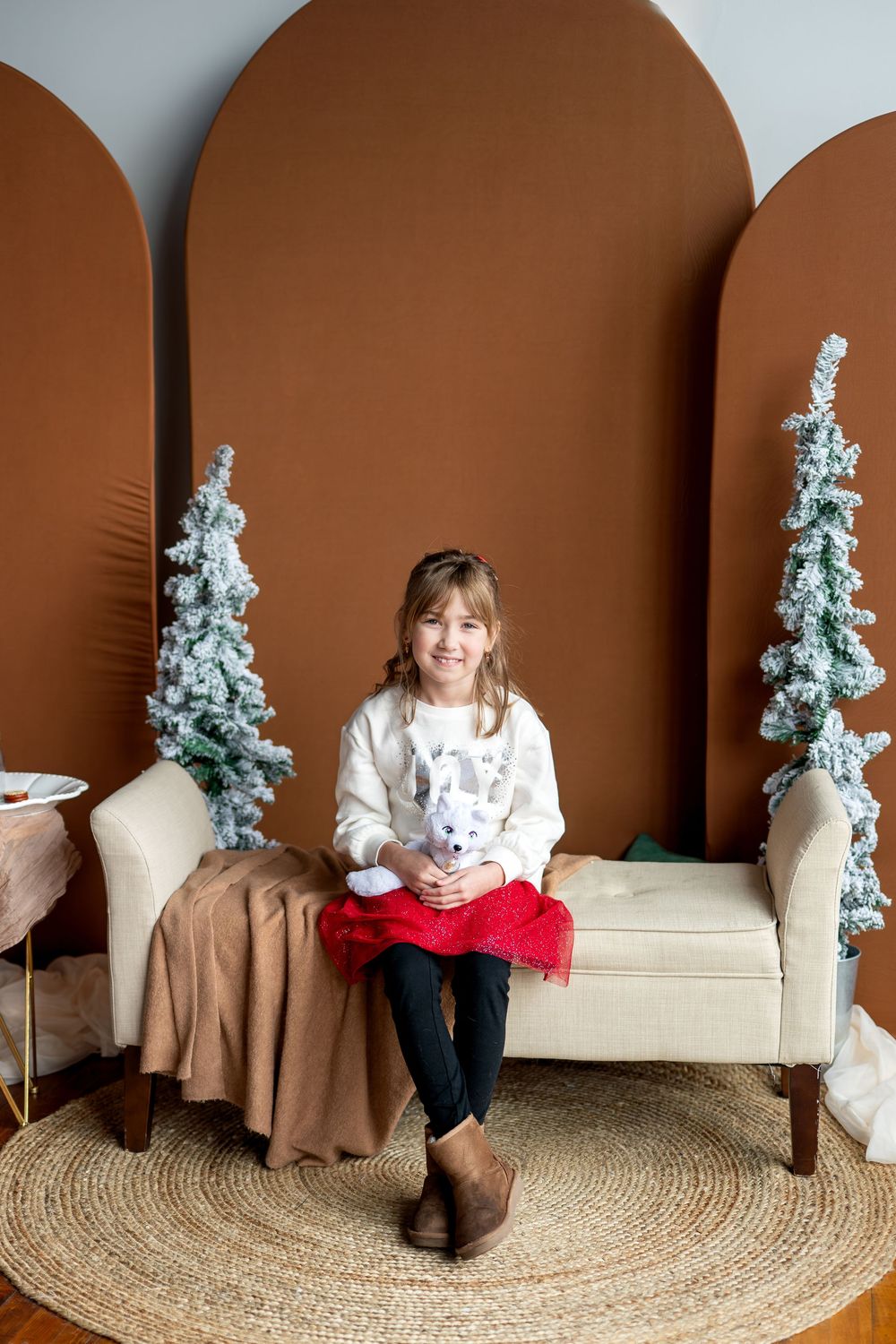 Young girl in white sweater and red skirt sits on cream bench with stuffed animal amid snowy Christmas trees.