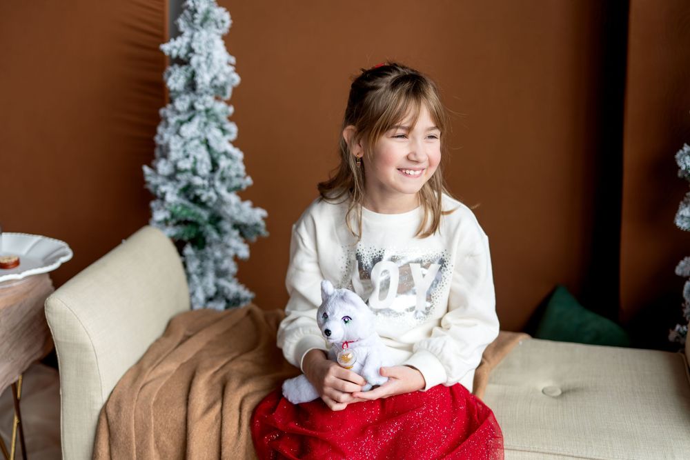 Smiling girl in white sweater holds stuffed unicorn toy while sitting on couch with Christmas trees behind her.