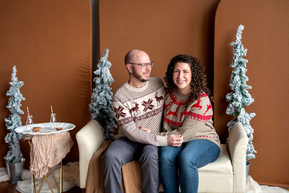 Smiling couple in coordinating reindeer holiday sweaters pose together on bench with snowy Christmas trees.