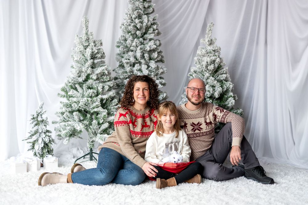 Family of three in holiday sweaters sit on white fluffy floor surrounded by snow-flocked Christmas trees.