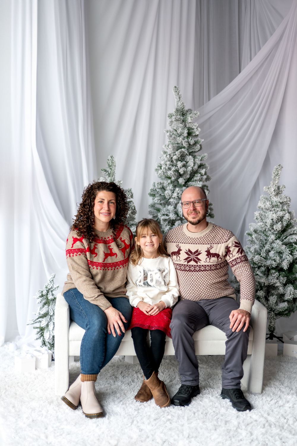 Family of three in Nordic sweaters sit together on white bench with snow-covered Christmas trees.
