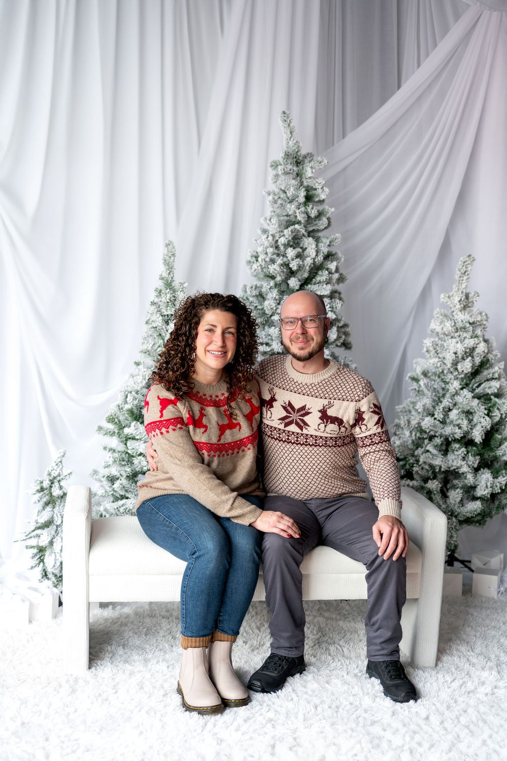 Couple in coordinating Nordic reindeer sweaters sit together on white bench by flocked Christmas trees.