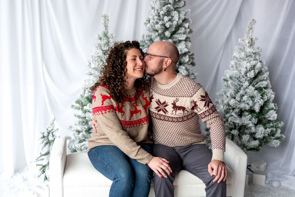 Couple in Nordic reindeer sweaters share a kiss on white bench surrounded by snowy Christmas trees.