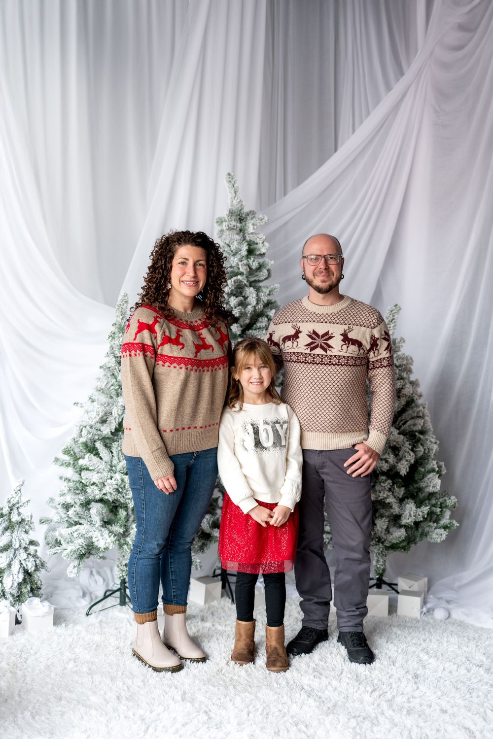 Family of three in holiday sweaters stand together before snow-flocked Christmas trees in white studio.
