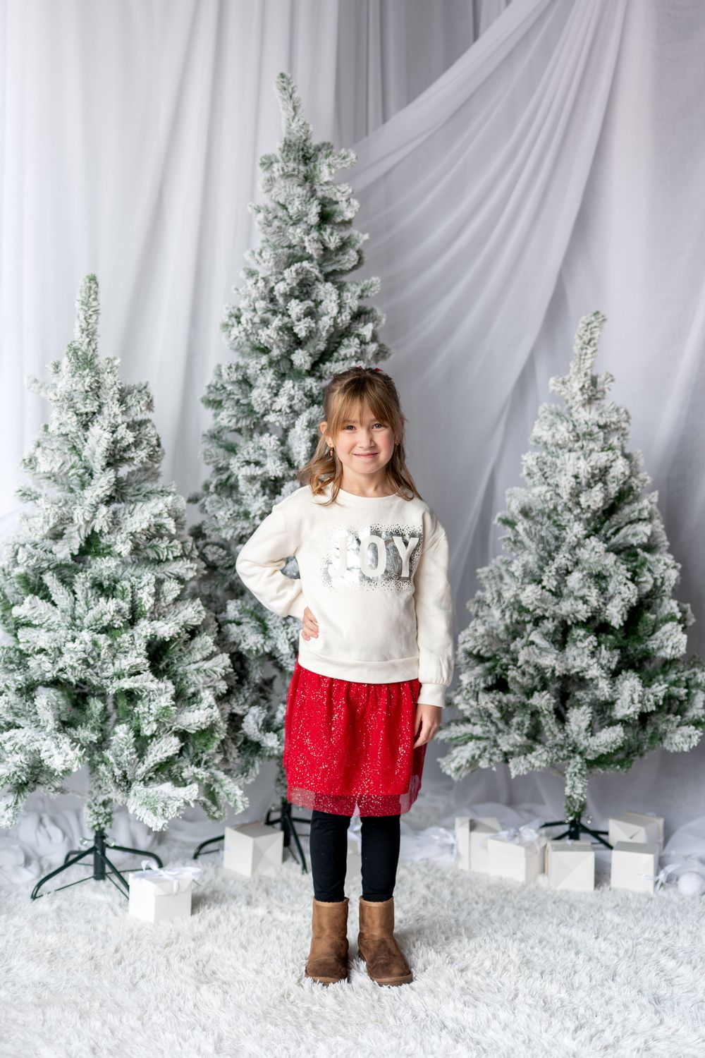 Young girl in JOY sweatshirt and red sequin skirt stands before snow-flocked Christmas trees with gifts.