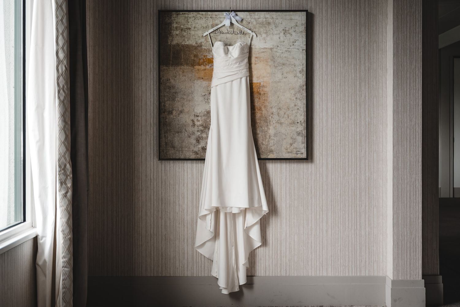 White wedding dress hanging elegantly against a neutral textured wall in natural light.