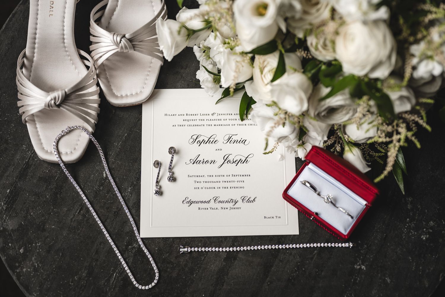 Wedding details flatlay with white flowers, invitation cards and bridal accessories on dark surface.