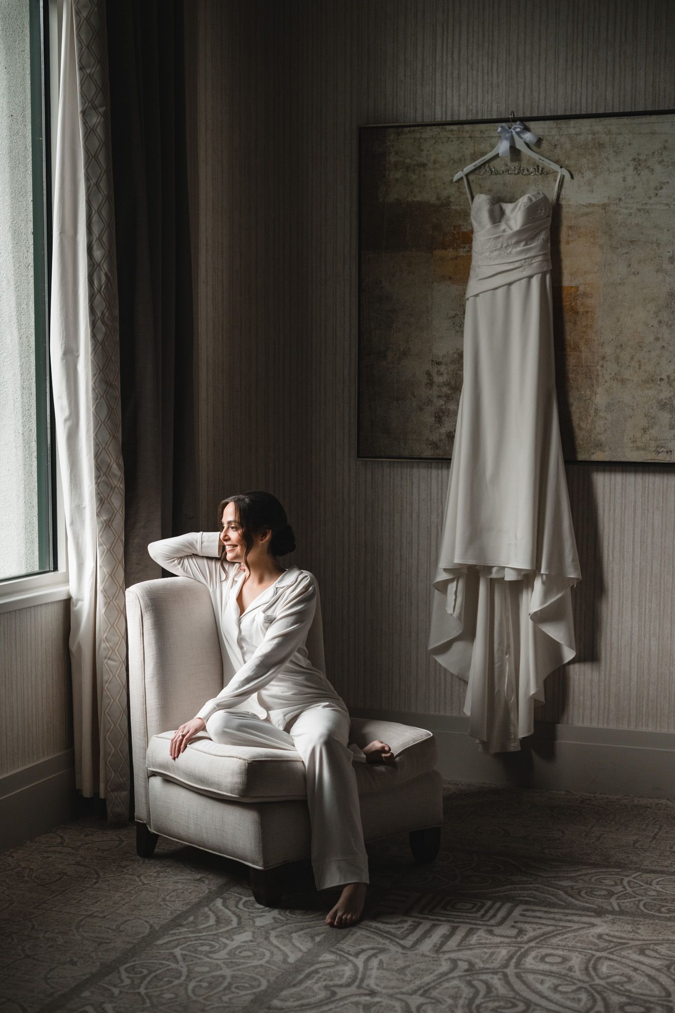 A bride in a white wedding gown sits elegantly in a room with white curtains and patterned carpet.