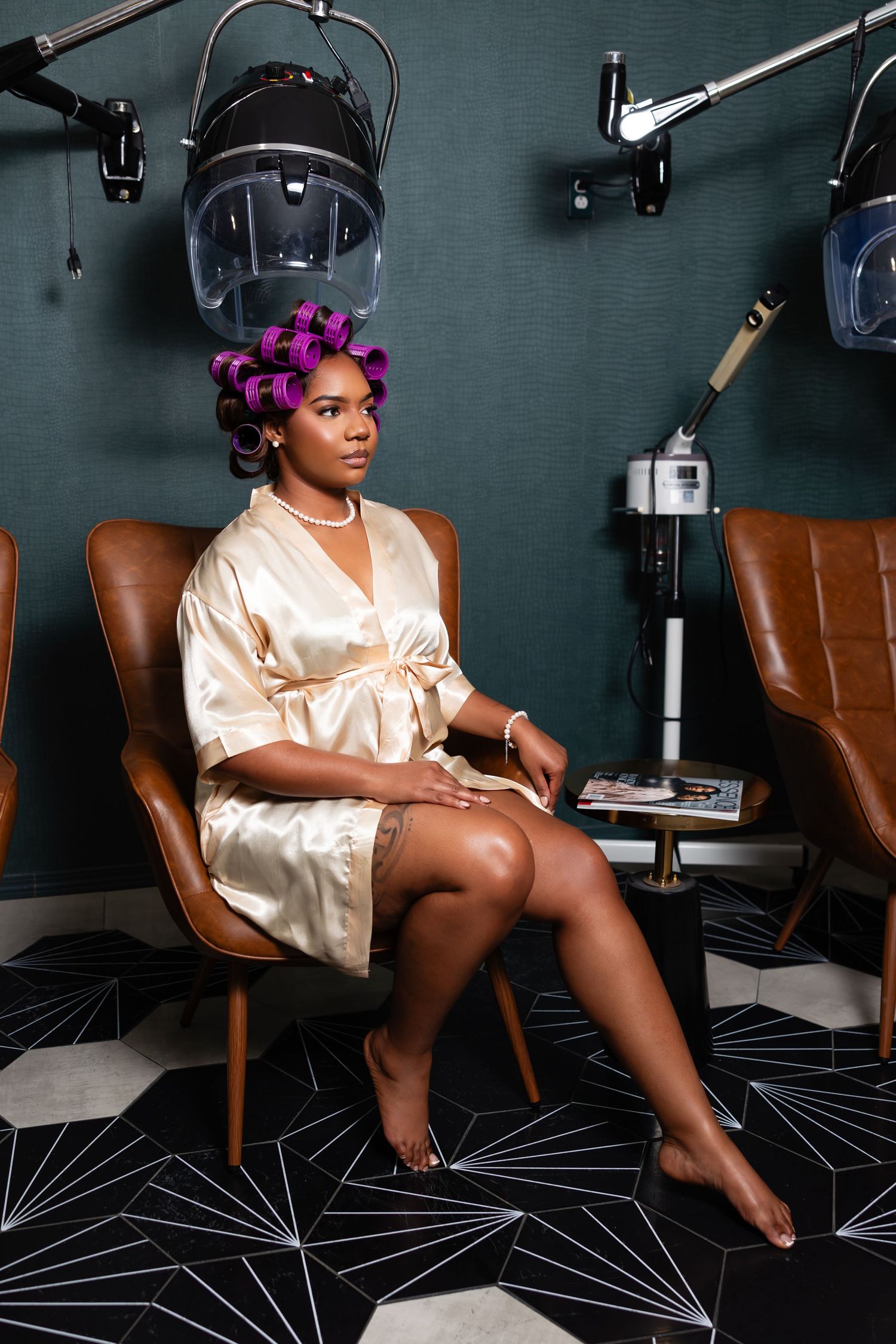 A person in a silk robe poses on brown leather chairs in a dark studio setting with beauty salon equipment.
