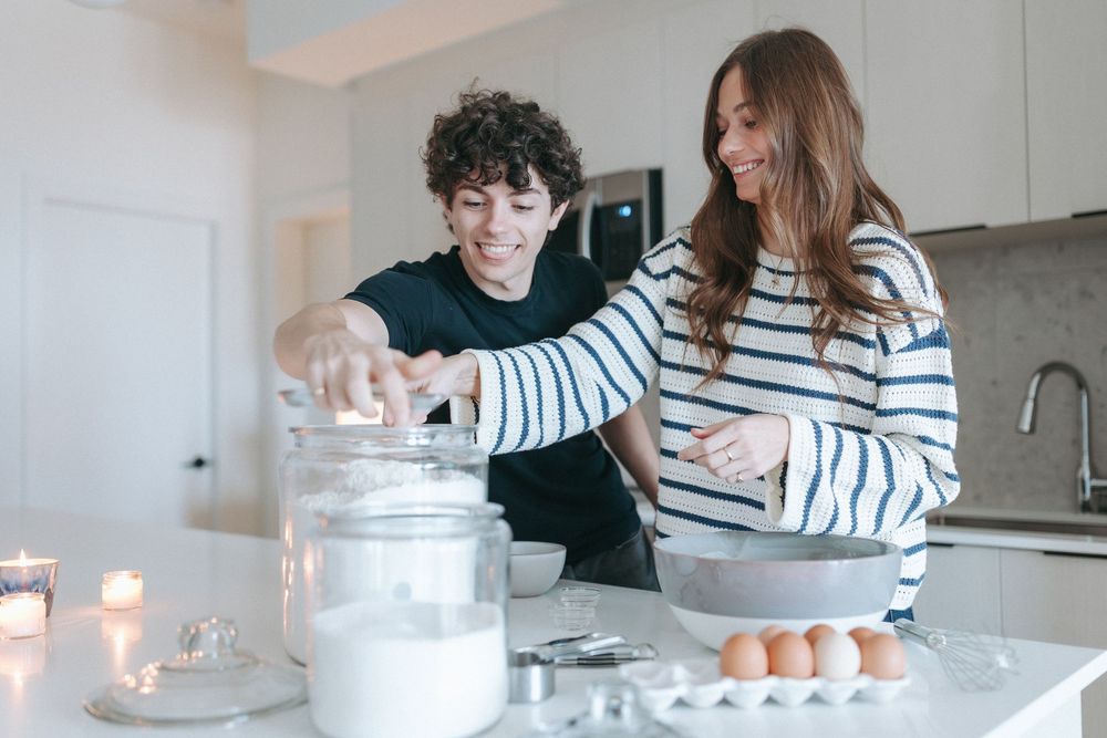 Couple working together in kitchen with baking ingredients and glass jars on counter.