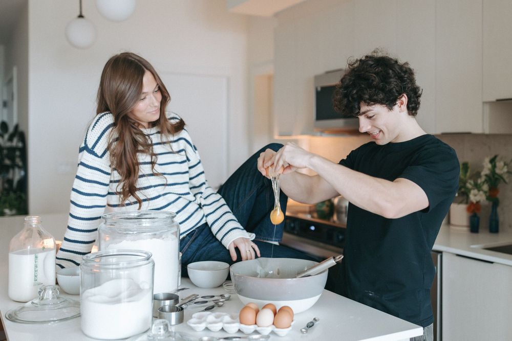 Couple preparing ingredients at kitchen counter with eggs and glass containers.