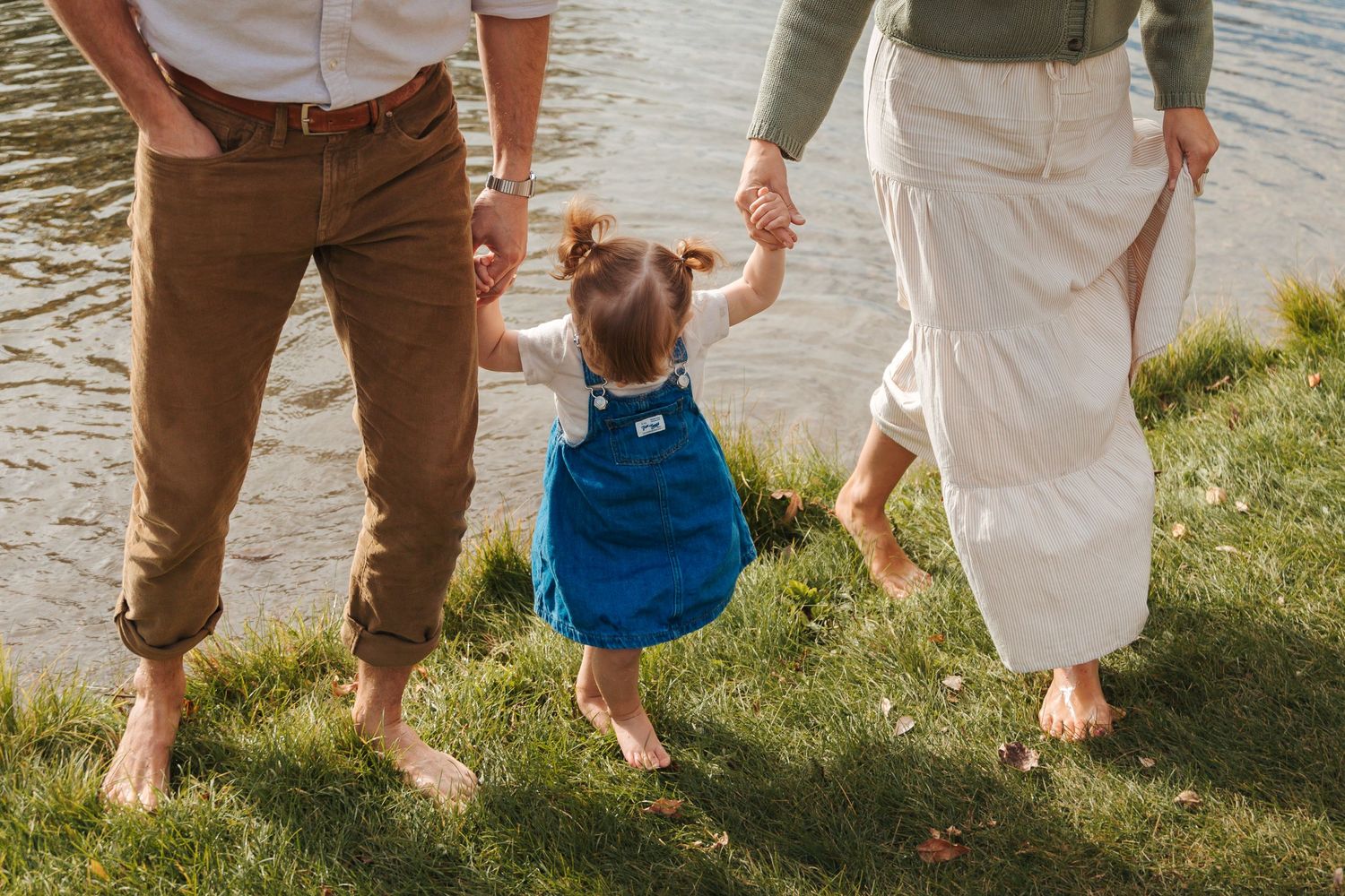 Parents hold hands with their toddler in blue overalls near a sunlit lake on green grass.
