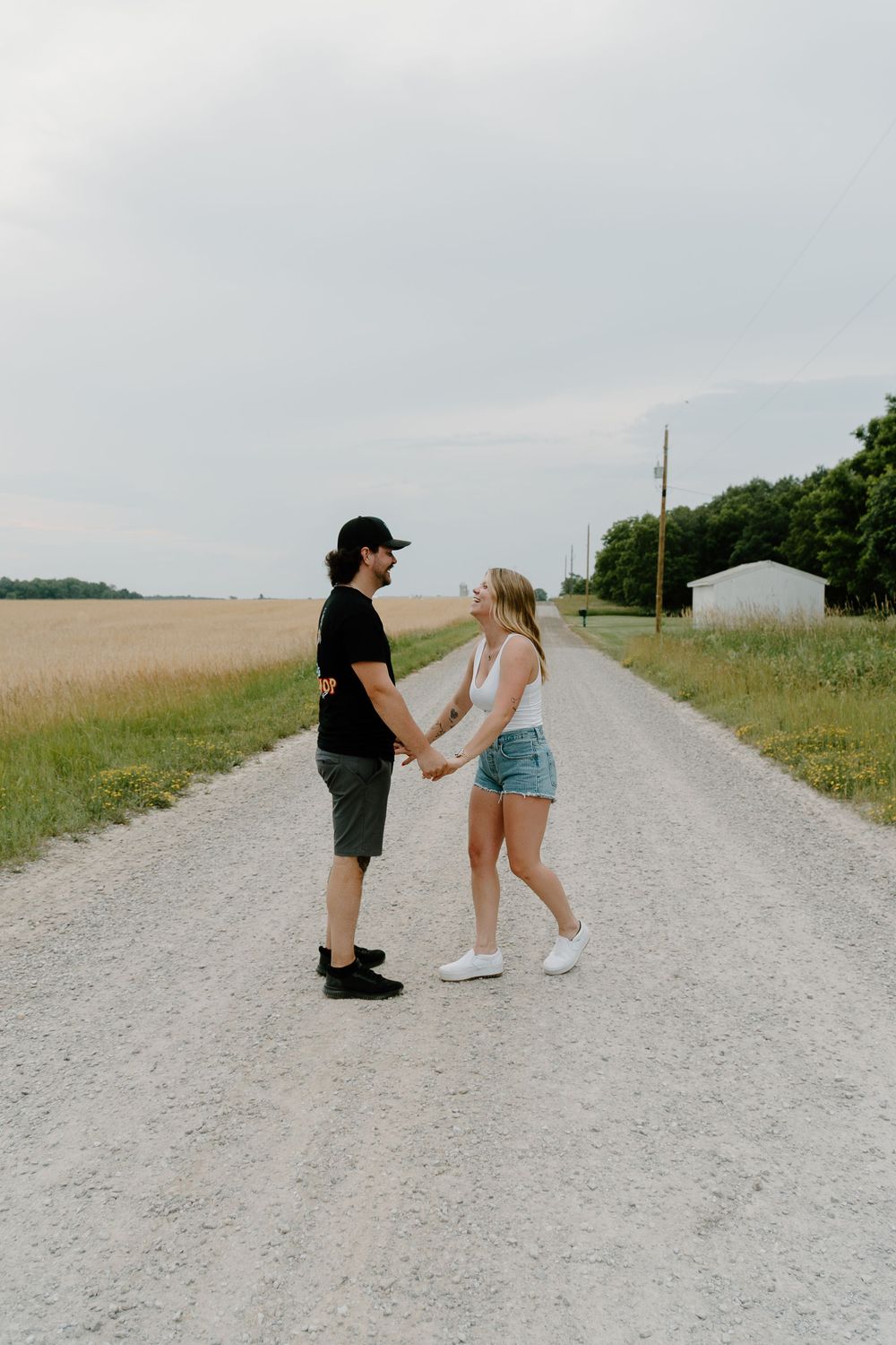 Two people interact on a gravel country road surrounded by grassy fields on a cloudy summer day.