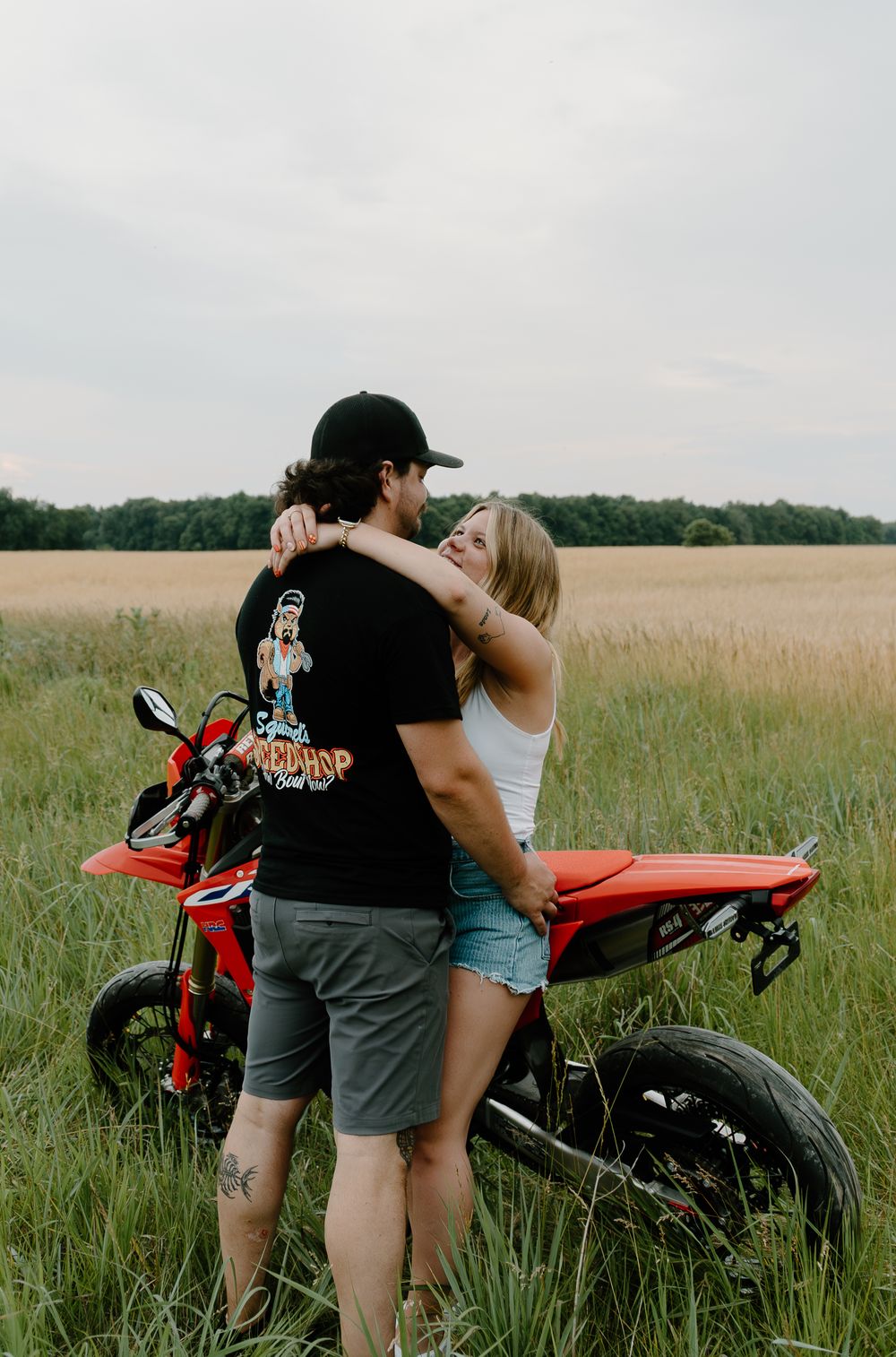 A couple shares an intimate moment beside a red motorcycle in a serene meadow at sunset.