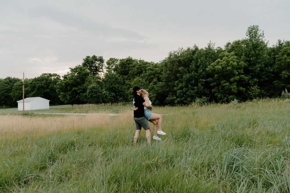 Two people walking through a tall grass meadow on a sunny summer day with trees in the background.