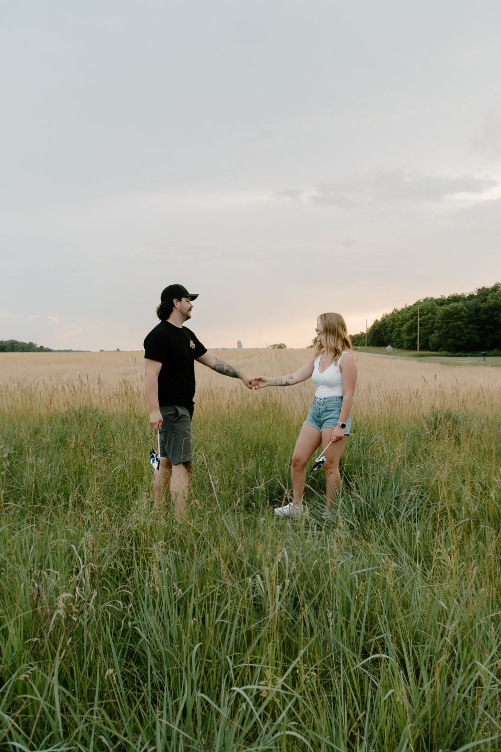 A couple stands together in a tall grass field during sunset, creating a romantic summer scene.
