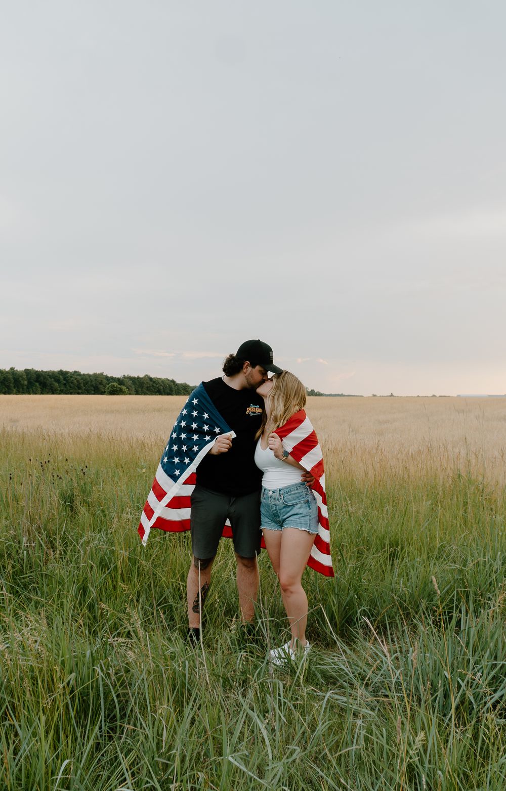 A couple draped in an American flag share a romantic moment in a grassy field at sunset.