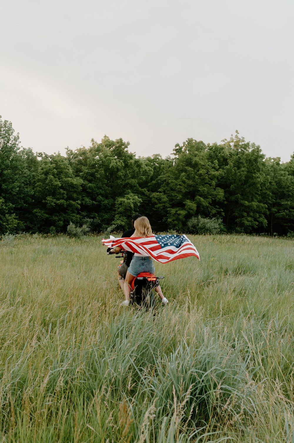 Person standing in tall grass field holding American flag against cloudy sky in patriotic outdoor scene.