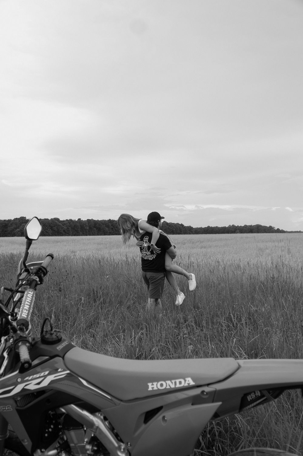 A red Honda dirt bike parked in a grassy field with someone examining it against a cloudy sky backdrop.