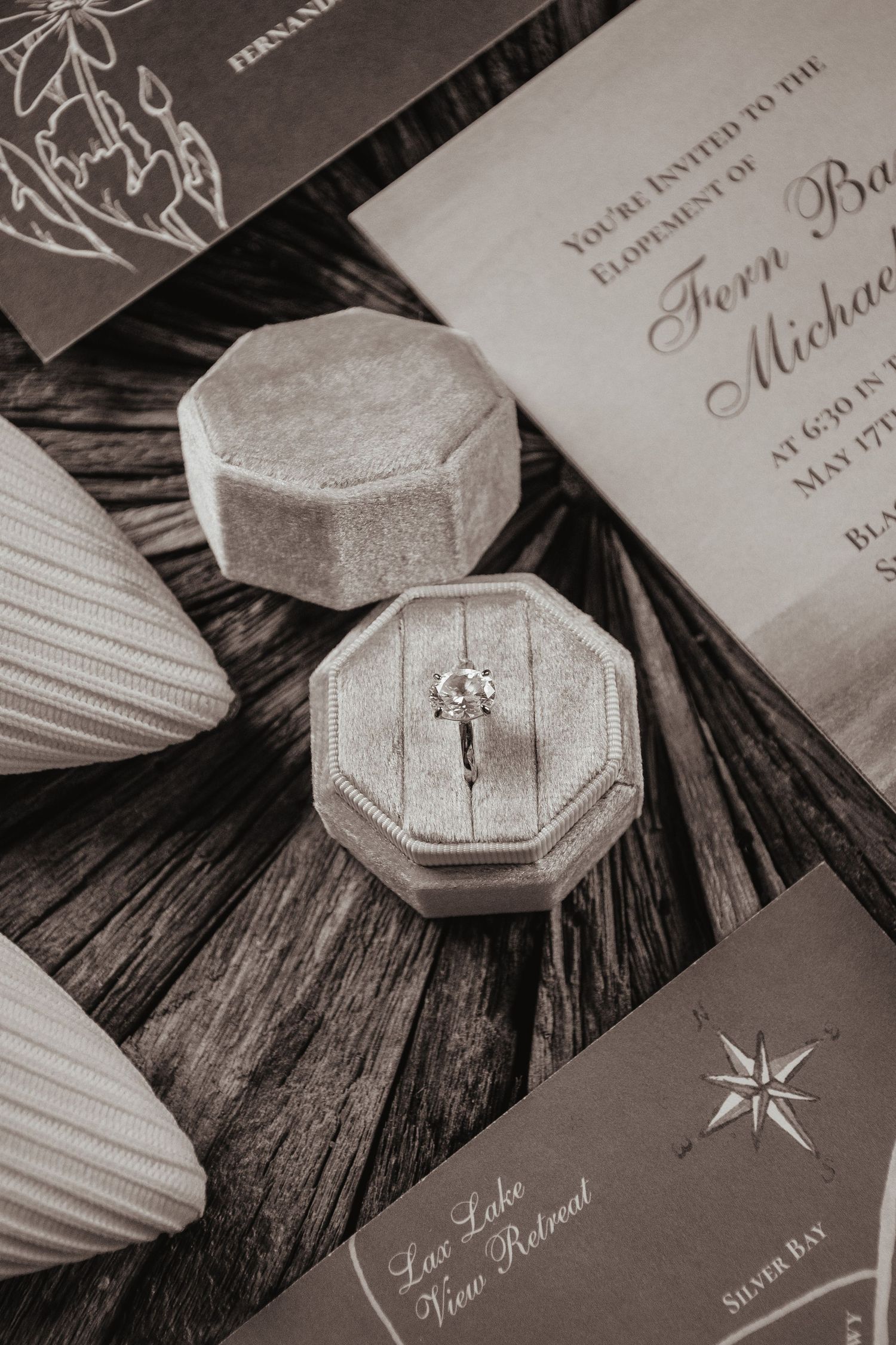 An engagement ring in a velvet hexagonal box sits among wedding invitation cards on a rustic wooden surface.