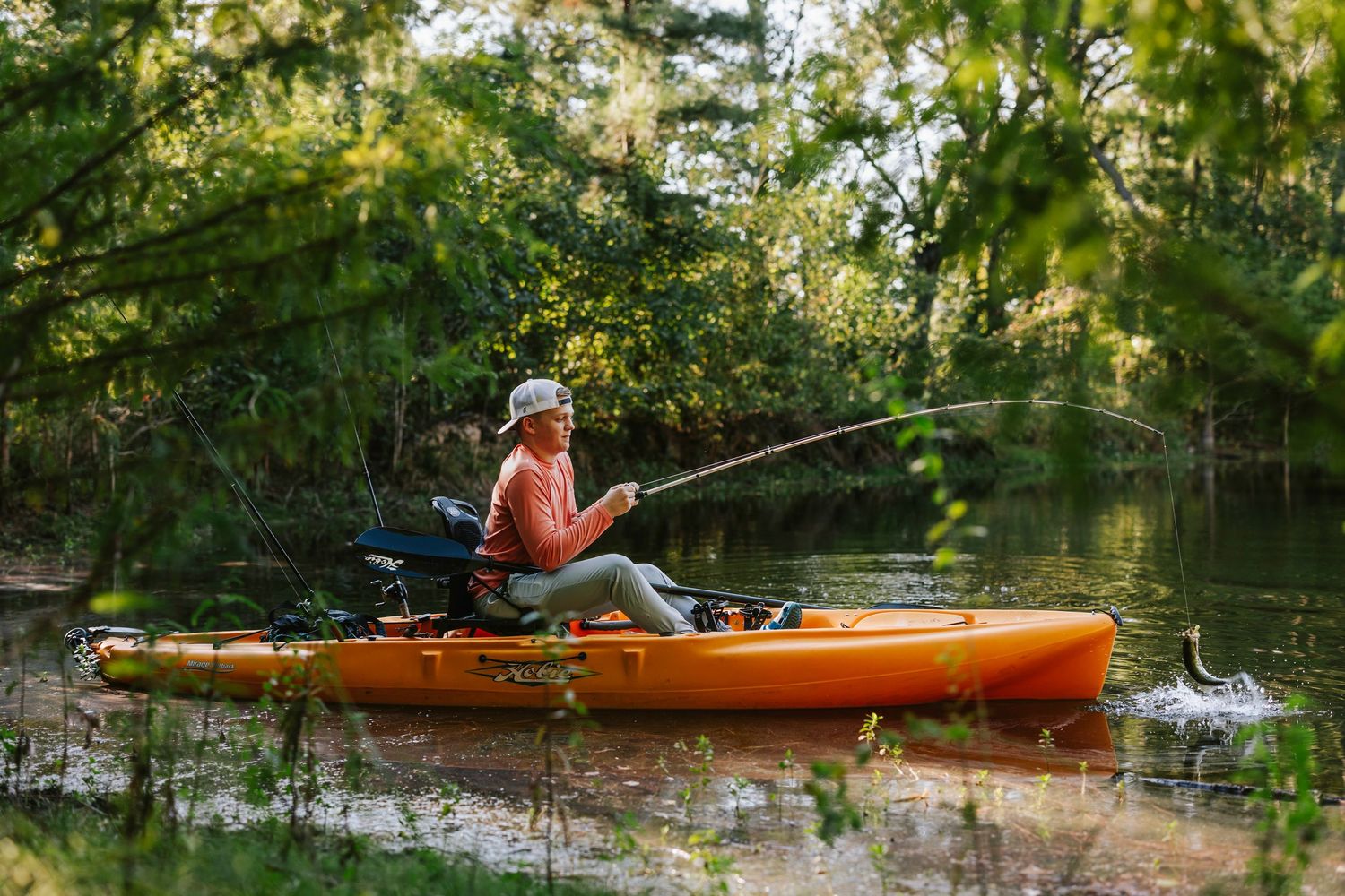 Person fishing from an orange kayak on a peaceful river surrounded by trees.