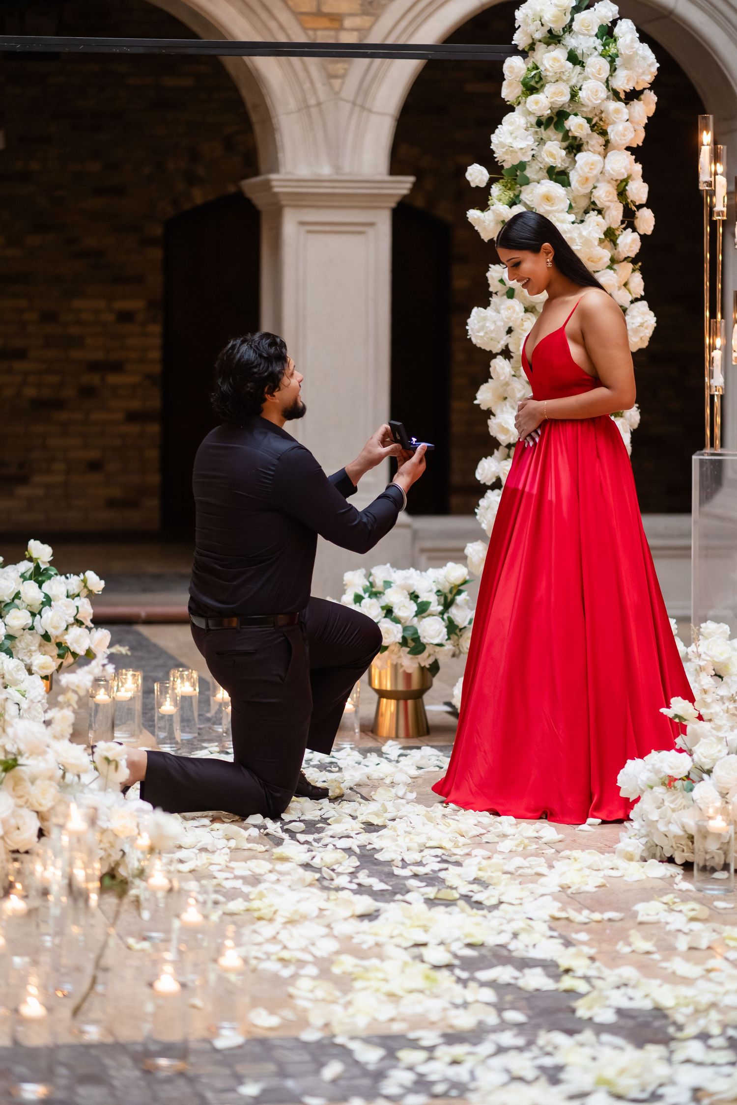 A romantic proposal scene with white floral decorations, candles and a person in a stunning red gown in an elegant archway.