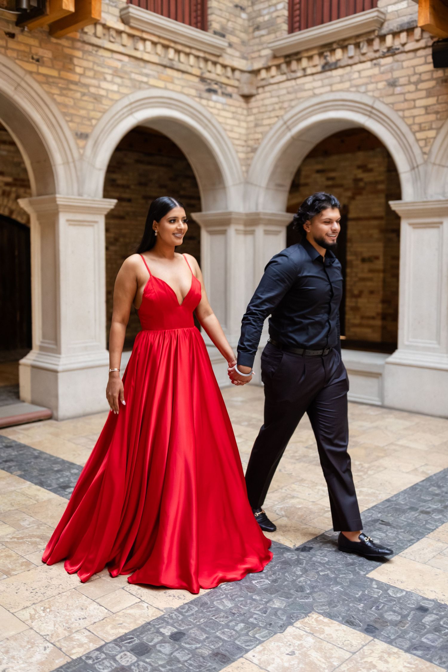 A couple poses in a historic courtyard with stone archways and brick walls, the woman wearing a flowing red dress.