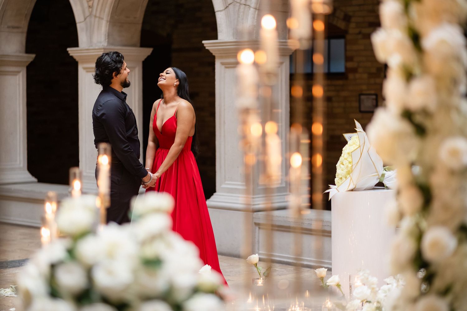 A couple in formal attire stands in an elegant archway surrounded by romantic candlelight and white floral decorations.