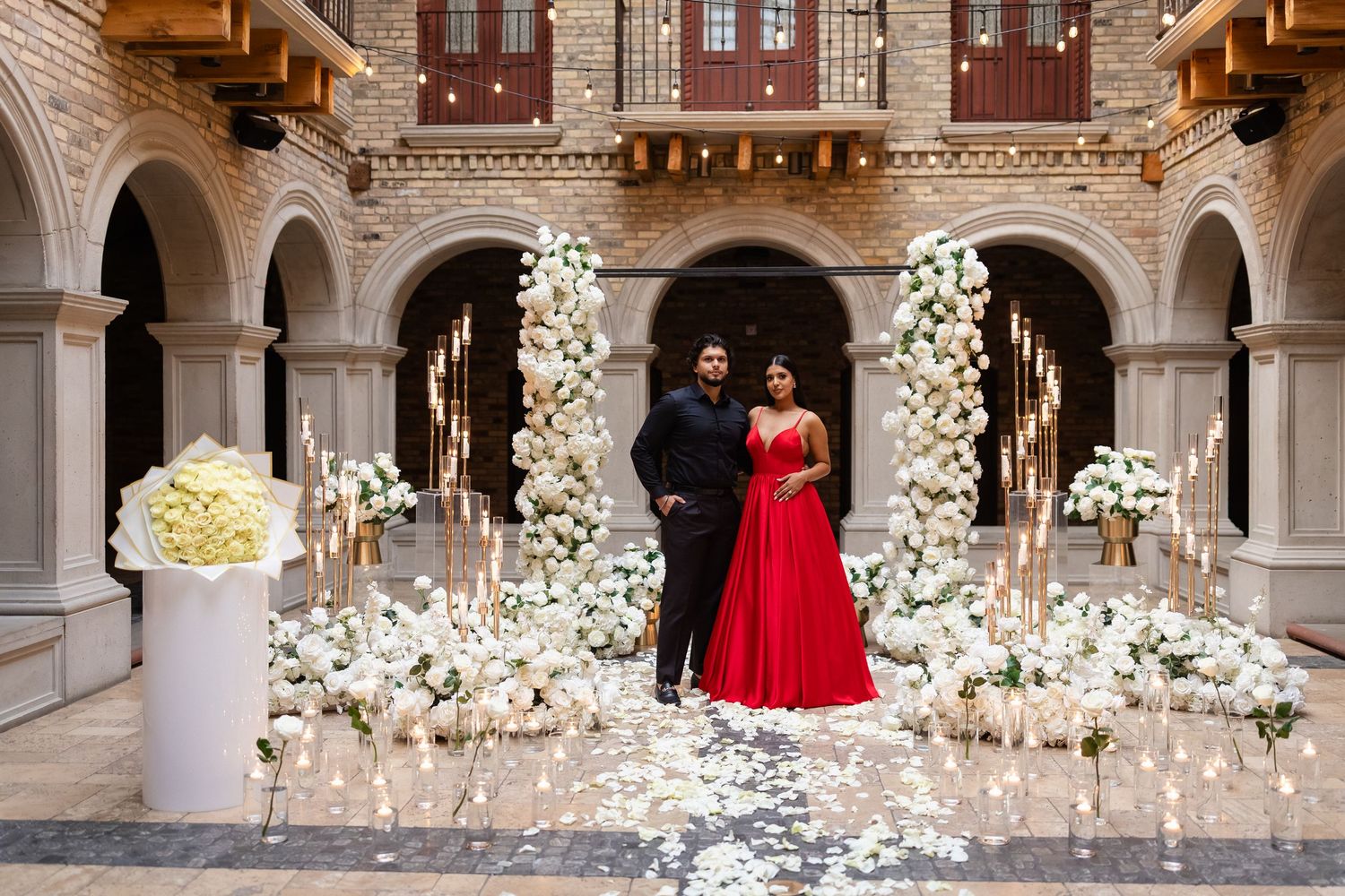 Red formal dress stands out against classical architecture adorned with white flowers and candlelight.
