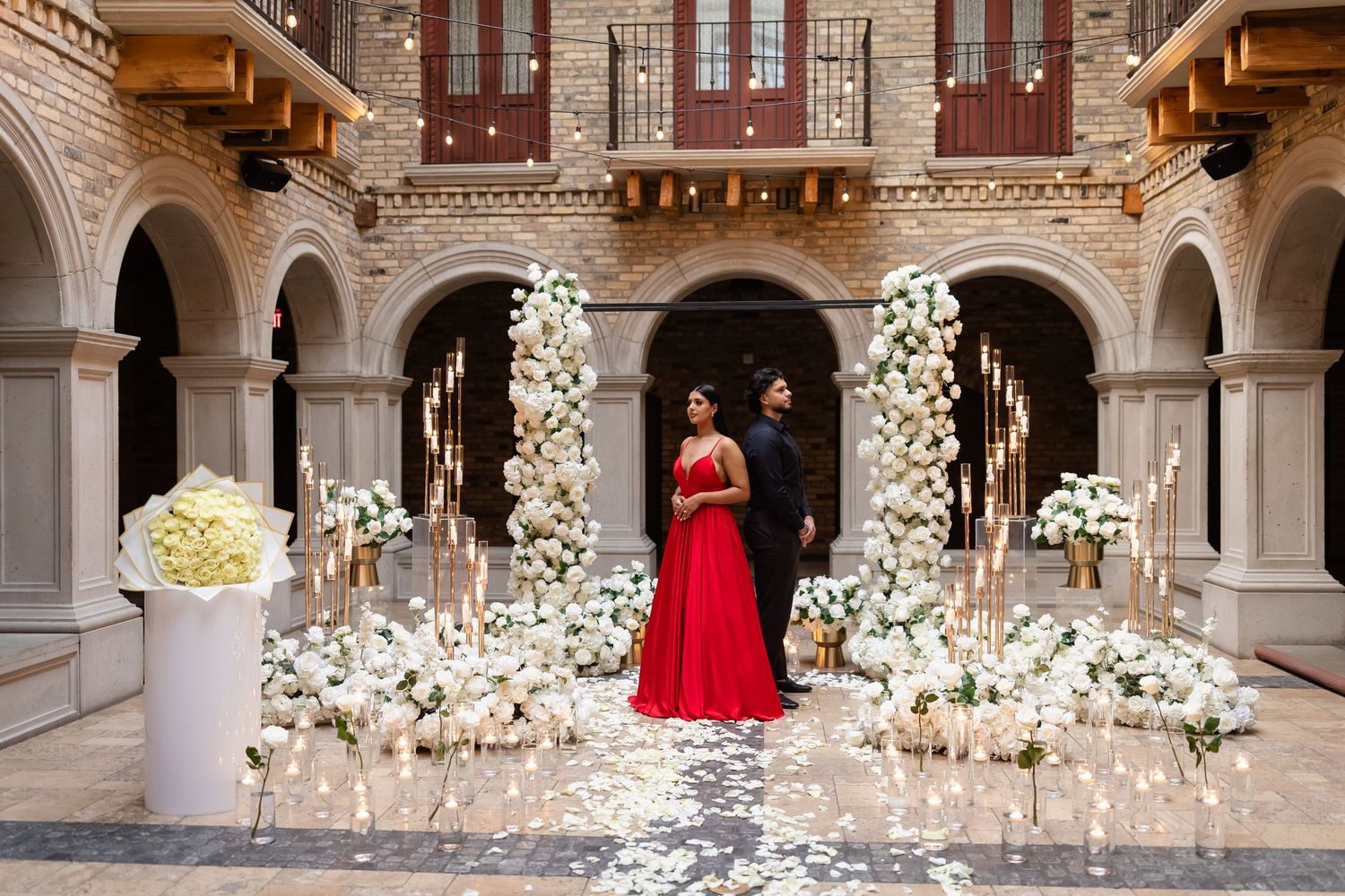 A bride in a red gown stands in an elegant marble hallway surrounded by glowing candles and white floral arrangements.