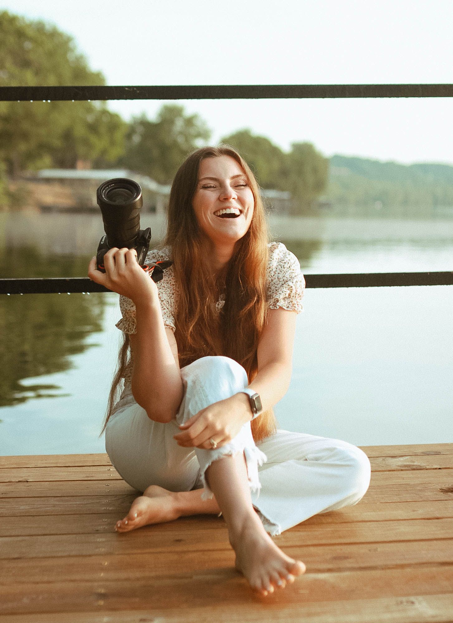 Person in white dress laughs joyfully while sitting barefoot on wooden dock by serene lake during sunset.