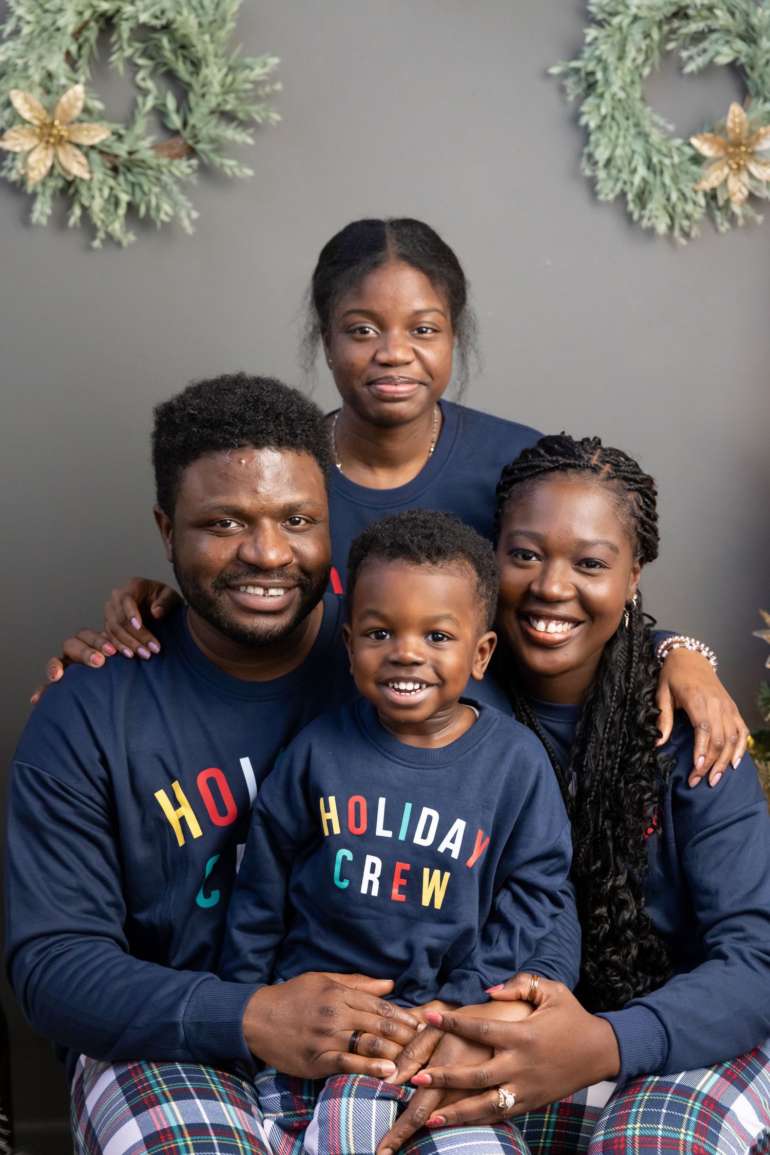 A family wearing matching holiday sweaters poses together against a gray background with festive wreaths.