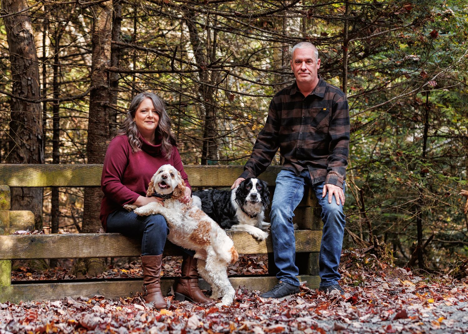 A couple sits on wooden steps in an autumn forest with their two dogs during a photo session.
