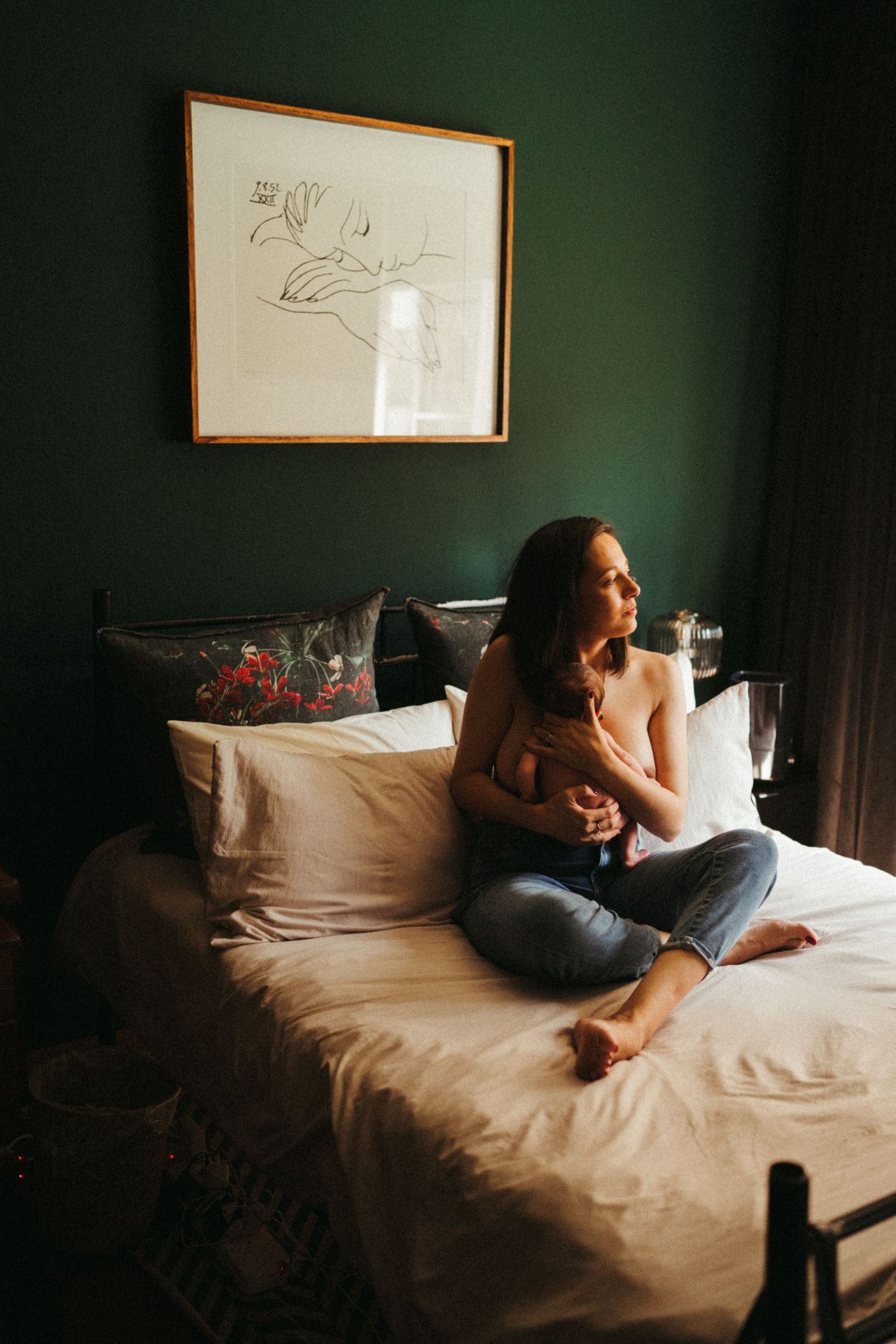 Woman lying on white bedding in moody dark room with artwork on green wall.