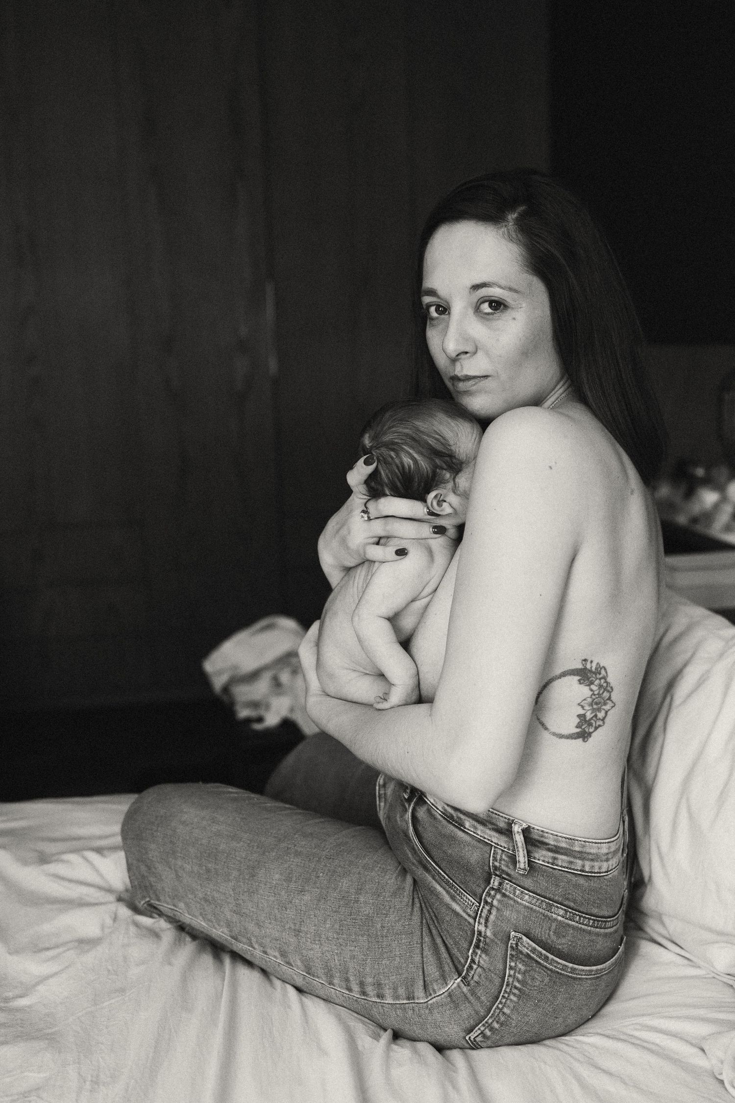 Woman in denim resting with newborn on white bedding.