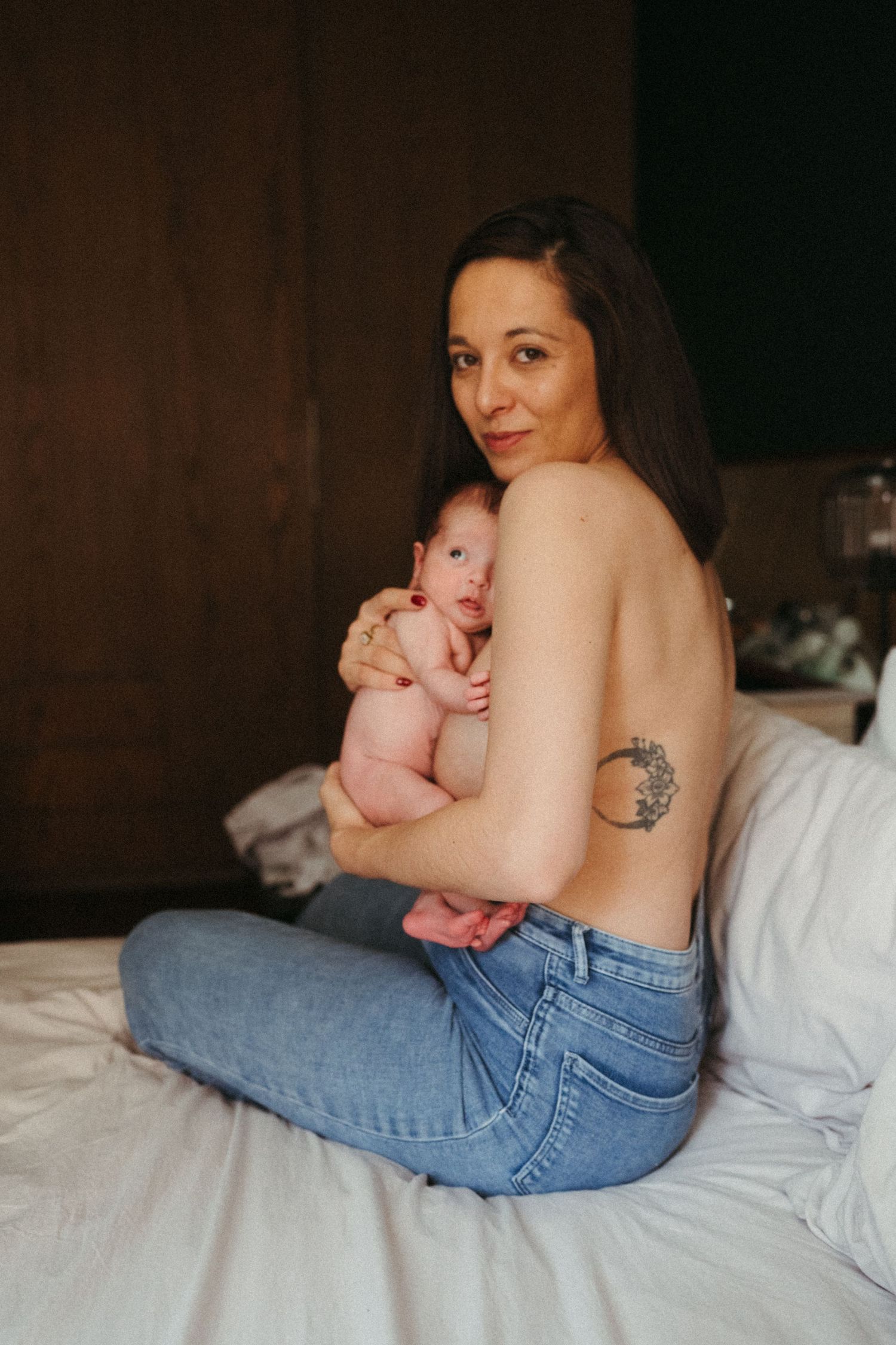 Mother in jeans holding newborn baby while lying on white bed.