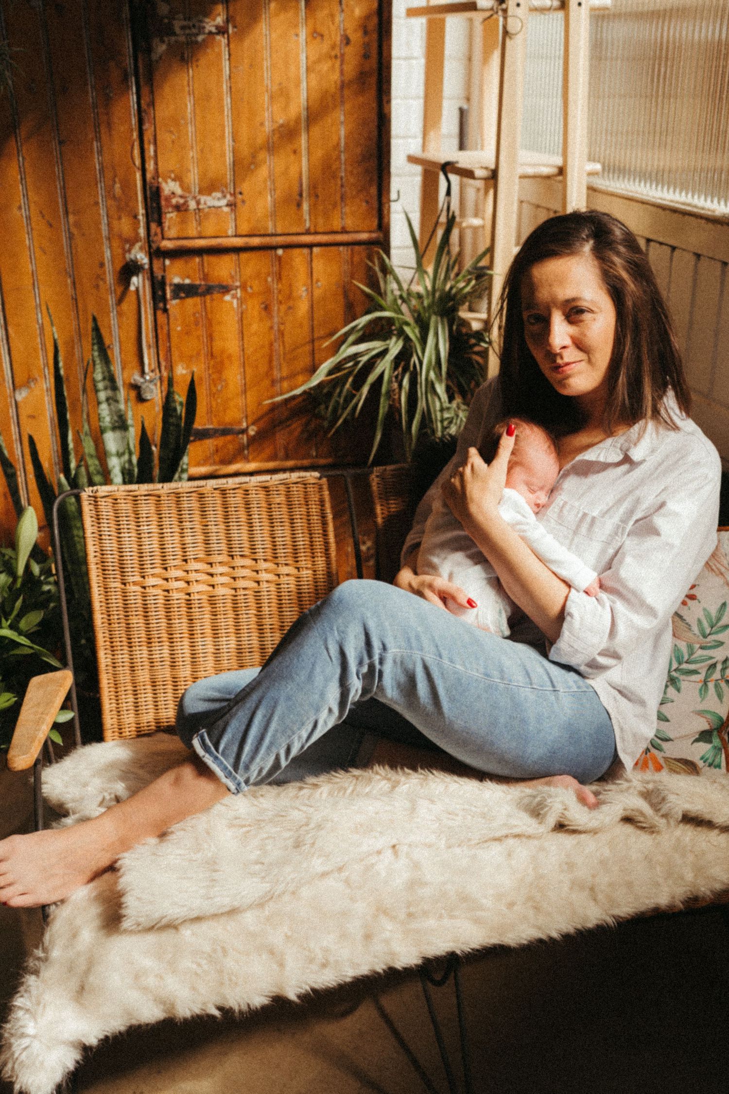 Woman resting with baby on furry white rug against wooden wall with plants.