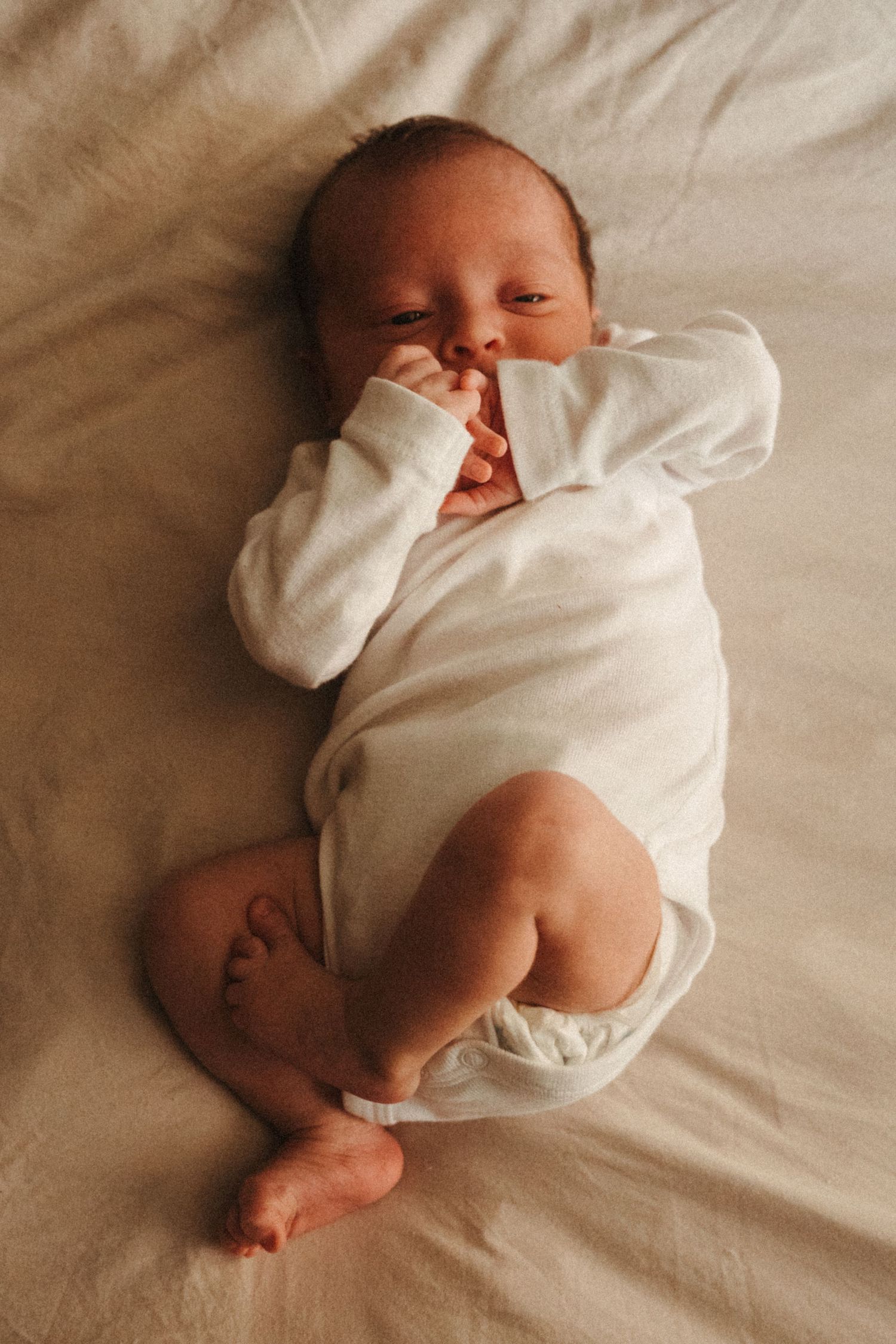Newborn baby in white onesie sleeping peacefully on light colored bedding.