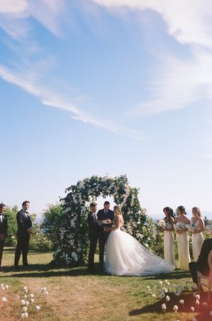 Outdoor wedding ceremony setup with floral arch and guests gathered on a hilltop under blue skies.
