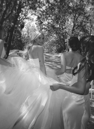 Wedding party walking along tree-lined path in flowing white dresses.